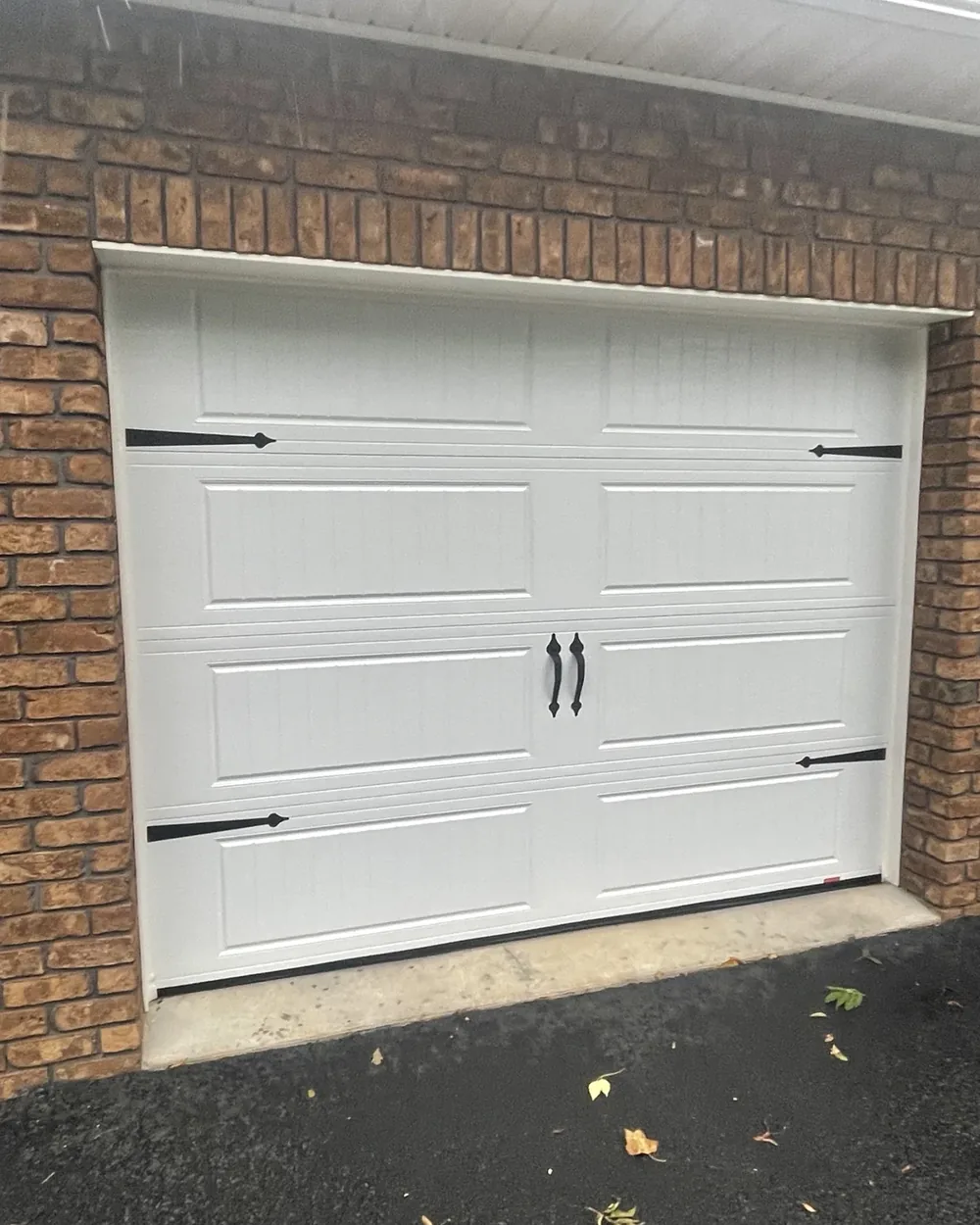 White carriage-style residential garage door with black hardware set in a brick wall